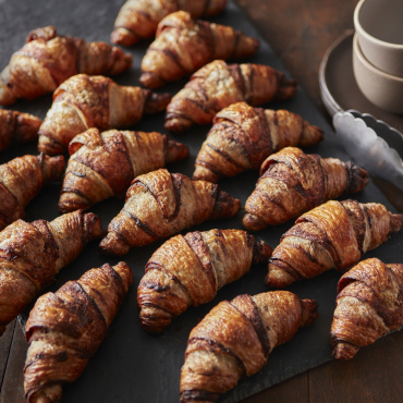 Chocolate croissants on a serving tray with tongs and coffee cup in the background